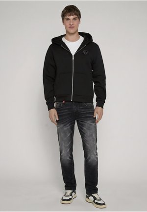 Junger Mann steht und trägt einen schwarzen Hoodie mit Reißverschluss, ein weißes T-Shirt, ausgewaschene schwarze Jeans und schwarz-weiße Turnschuhe vor einem schlichten Hintergrund.