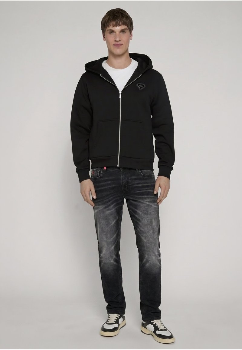 Jeune homme debout portant un sweat à capuche zippé noir, un t-shirt blanc, un jean noir délavé et des baskets noir et blanc sur un fond uni.
