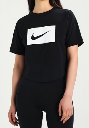 Personne portant un t-shirt noir avec un logo rectangulaire blanc arborant une virgule noire Nike sur la poitrine, assorti d’un pantalon noir.