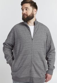 Felpa grigia con zip in un tessuto morbido e texturizzato. Collo alto, maniche lunghe, tasche laterali e vestibilità rilassata. T-shirt bianca sotto.