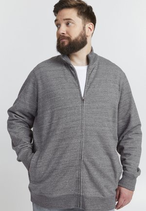 Sudadera gris con cremallera en un tejido suave y texturizado. Cuello alto, mangas largas, bolsillos laterales y ajuste holgado. Camiseta blanca debajo.