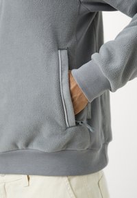 Γκρι φούτερ με γούνα που διαθέτει τσέπη με φερμουάρ στο πλάι, ριμπ στα μανικιά και υφή υφάσματος. Λείο φινίρισμα με casual σχεδίαση.