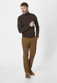Dunkelbrauner Rollkragenpullover, eng anliegend. Khakifarbene Chinos mit sichtbaren Taschen und Nähten. Braune Schnürschuhe. Neutraler Hintergrund.