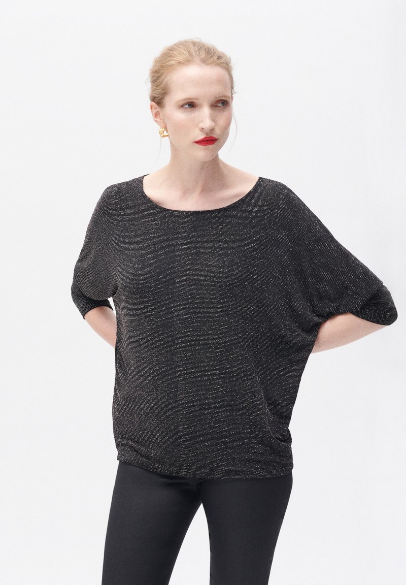 Schwarzes Stricktop mit lockerer Passform, weitem Ausschnitt und kurzen Ärmeln. Das Material hat einen dezenten Glitzereffekt. Getragen mit schwarzen Hosen.