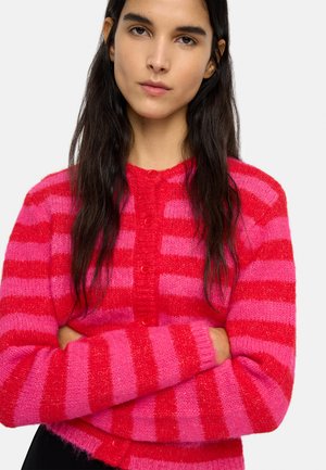 Młoda kobieta o długich ciemnych włosach, ubrana w czerwony i różowy pasiasty, miękki, zapinany na guziki kardigan, z założonymi ramionami.