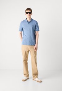 Tricou polo de culoare bleu deschis cu un finisaj texturat, pantaloni bej cu croială dreaptă și adidași albi cu accente bej. Modelul poartă ochelari de soare închiși la culoare.