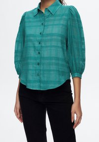 Mujer con blusa transparente de cuadros color verde azulado con mangas abullonadas hasta el codo y pantalones negros, de pie con los brazos relajados a los lados.