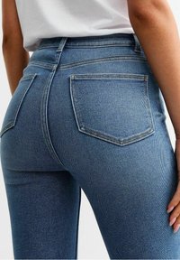 Persona que lleva puestos unos pantalones ajustados de cintura alta azules y una camisa blanca, con los bolsillos traseros visibles y la mano descansando en el muslo.