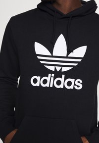Svart huvtröja med en vit Adidas-logotyp och tre ränder, med en framlomma och snörning i huvan. Tillverkad av mjukt material.