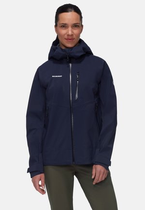 Kobieta stoi, ubrana w granatową kurtkę z kapturem na zamek błyskawiczny, z kieszeniami na piersi i bokach, w połączeniu z oliwkowymi spodniami.