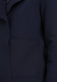 Ciemnogranatowa kieszeń płaszcza z przeszytym detalem logo na materiale, z widoczną częścią rękawa i otwarciem z przodu.