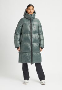 Gewatteerde lange jas in glanzend donkergroen met een hoge kraag, ritssluiting aan de voorkant en een oversized silhouet. Bevat elastische manchetten en zijzakken.