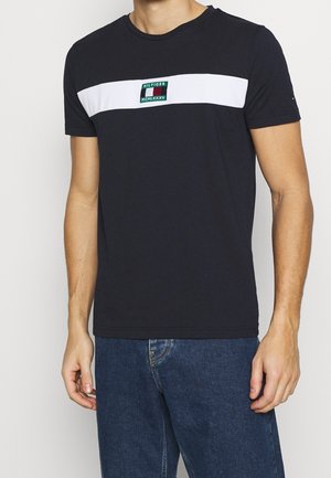 Mężczyzna noszący czarną koszulkę z białym poziomym paskiem i małym zielonym, czarnym i białym logo na klatce piersiowej, w połączeniu z niebieskimi dżinsami.