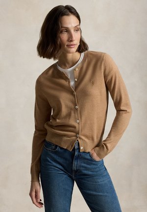 Femme aux cheveux foncés mi-longs, portant un cardigan camel boutonné sur une chemise blanche et un jean bleu, debout avec une main dans la poche.