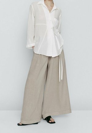 Pantalon large beige ample associé à un chemisier blanc boutonné style cache-cœur avec des sandales noires à bouts ouverts sur un fond neutre.