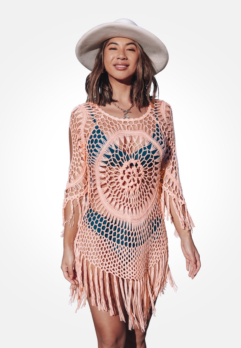 Szydełkowy nakrycie w miękkim odcieniu brzoskwiniowym z okrągłym wzorem, z frędzlami na dolnym brzegu i rękawami o długości trzy czwarte, założone na niebieski strój kąpielowy.