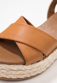 Tan synthetisch leren wedge sandaal met gekruiste banden, geweven jute zool en een gladde, matte afwerking op de voetbed.