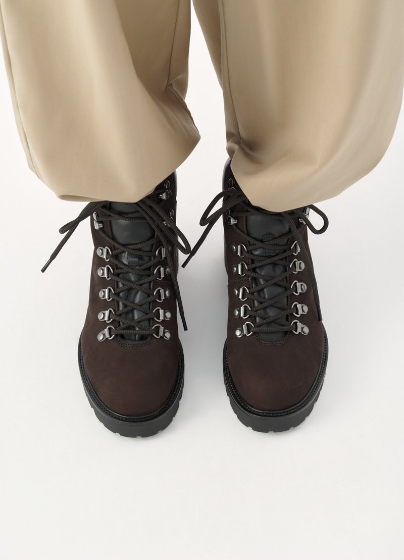 Bruine wandelschoenen met een bovenwerk van suède, metalen ogen en zwarte veters. Dikke rubberen zolen zorgen voor grip. Beige broeken zichtbaar erboven.