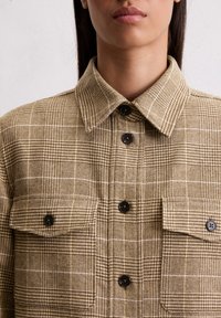 Chemise à carreaux dans des tons beige et marron, avec un col pointu, deux poches poitrine et des boutons noirs. Tissu texturé avec un motif en quadrillage.