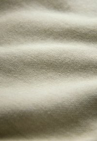 Close-up van lichtbeige stof met een fijne geweven textuur en zachte plooien die subtiele schaduwen en highlights creëren.