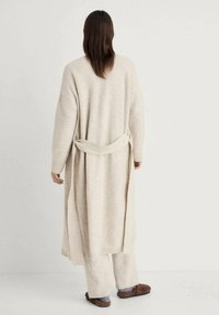 Cardigan lungo beige chiaro con cintura da annodare, texture a maglia, maniche lunghe e schiena aperta. Abbinato a pantaloni beige larghi e pantofole marroni.