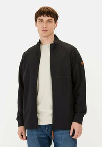 Zwarte zip-up jack met een hoge kraag, gemaakt van textuurstof, met een oranje lederen tabaccent en ribbelmanchetten. Gedragen over een licht shirt.