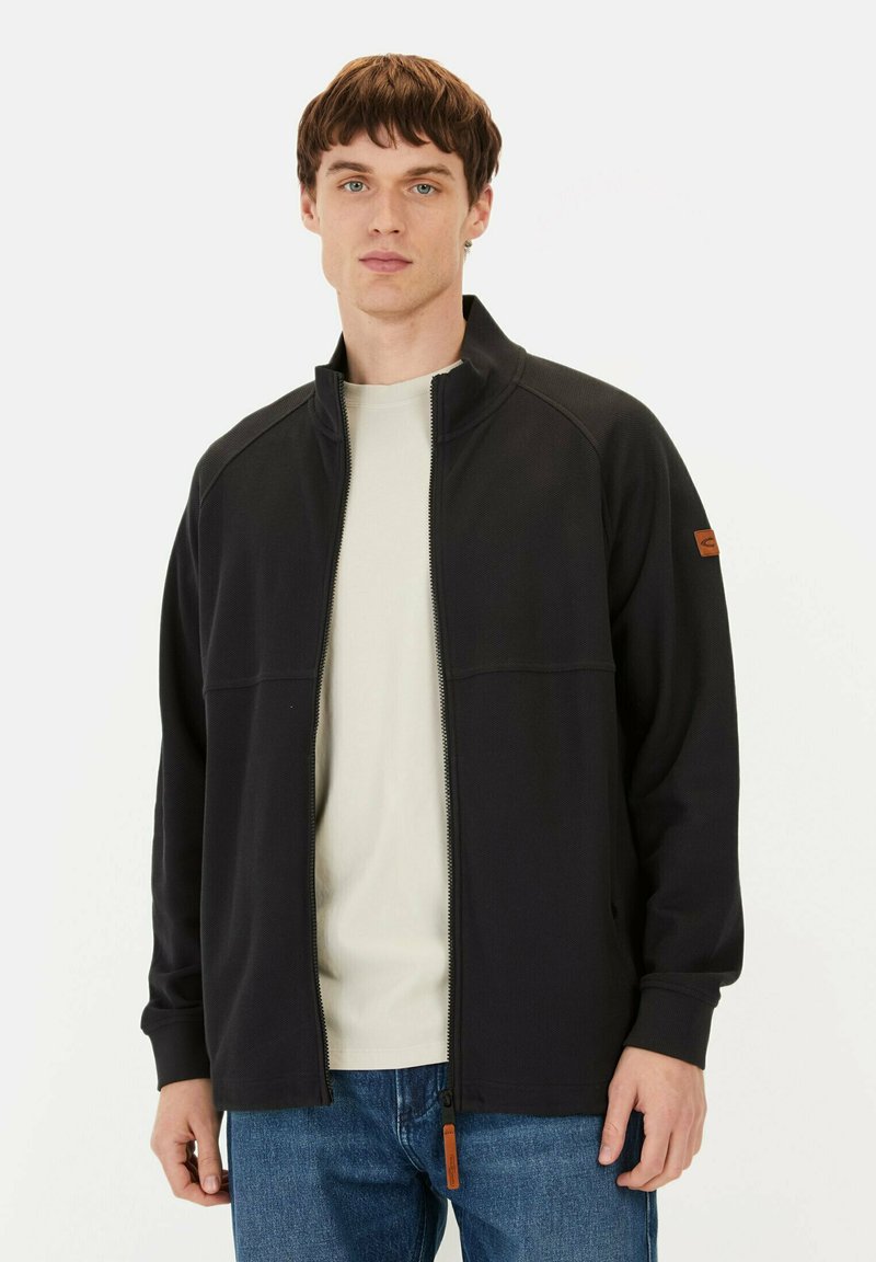 Zwarte zip-up jack met een hoge kraag, gemaakt van textuurstof, met een oranje lederen tabaccent en ribbelmanchetten. Gedragen over een licht shirt.