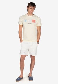 Camiseta de algodón de color amarillo claro con letras gráficas "H," "N," y un ancla en azul y rojo. Pantalones cortos blancos con patrones de anclas multicolores. Zapatillas azules.