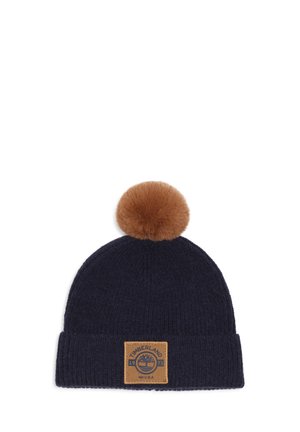 Bonnet en maille bleu marine avec un pompon en fourrure marron sur le dessus et un patch logo Timberland beige sur le revers.