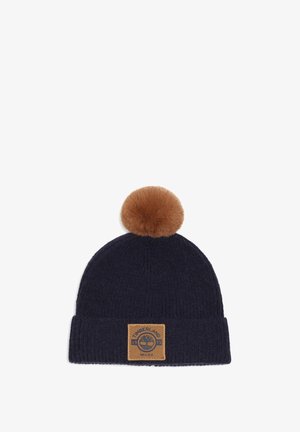 Donkerblauwe gebreide muts met een bruine bontpompon bovenop en een beige Timberland-logo patch op de boord.