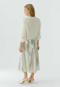 Vrouw in een zacht crèmekleurig trui, zilveren midi-rok met pailletten en crèmekleurige slingback-hakken, staand en naar links kijkend tegen een effen achtergrond.