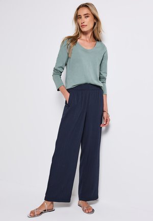 Hellgrünes Langarmoberteil kombiniert mit navyblauen, weit geschnittenen, plissierten Hosen, die einen lockeren Fit und seitliche Taschen bieten. Metallische Sandalen runden den Look ab.
