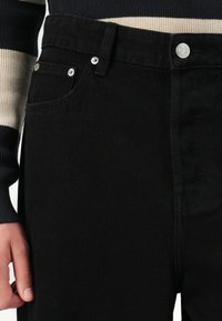 Person, der schwarze Denim-Jeans mit silbernen Nieten und Knöpfen trägt, kombiniert mit einem schwarz-beige gestreiften Langarmoberteil.
