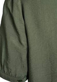 Vêtement en lin noir avec une finition douce et texturée. Il possède des manches froncées, un détail de bouton au niveau du poignet et une coupe décontractée.