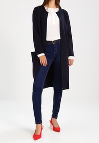 Marineblauer langer Cardigan mit offenem Vorderteil, weißem Langarmshirt, dunklen Jeans und roten High Heels. Aus glattem Stoff und minimalistischer Gestaltung.