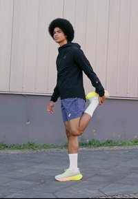 Chaqueta ligera negra, pantalones cortos azules, calcetines blancos y zapatillas deportivas con acentos neón. La persona estira una pierna mientras está de pie sobre el pavimento.