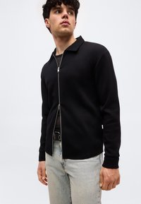 Νέος άνδρας που φοράει μαύρο πουλόβερ με φερμουάρ πάνω από μαύρο πουκάμισο και ανοιχτόχρωμα τζιν, κοιτάζοντας ελαφρώς στο πλάι, με φόντο απλό.