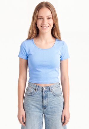 Ung kvinde med langt lysebrunt hår, iført ringformede øreringe, en lyseblå kortærmet crop top og lyseblå højtaljede jeans, smilende.