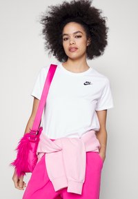 T-shirt en coton blanc avec un logo Nike noir, associé à un pantalon de survêtement rose. Un sac rose avec un accent en fourrure pend d'une épaule.