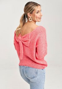 Oversized różowy sweter dzianinowy z V-kształtnym dekoltem z tyłu i detalem w postaci kokardy. Miękka tekstura, luźny krój, zestawiony z jasnoniebieskimi dżinsami.
