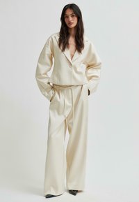 Kvinna iklädd lösa krämfärgade byxor och en matchande kort blazer med en enda knapp, stående mot en enkel ljus bakgrund.