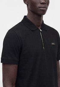 Homme portant un polo noir à manches courtes avec une fermeture éclair argentée à l'avant et un petit patch logo rectangulaire sur la poitrine.