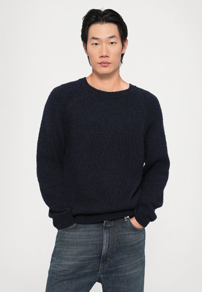 Suéter malhado azul-marinho com acabamento texturizado, decote redondo e punhos canelados; usado com jeans de ganga ajustados. Design simples, ajuste casual.