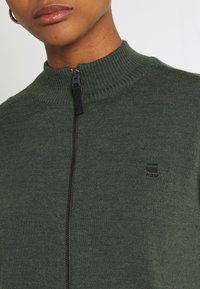 Donkergroene ritsvest met geribde kraag, zwarte rits en klein zwart logo op de borst. Zachte gebreide stof met een gestructureerde uitstraling.