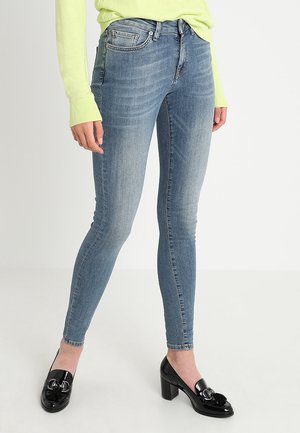 Jeans ajustados de color azul con un lavado desgastado, que presentan un diseño tradicional de cinco bolsillos y un corte slim. Combinados con tacones de charol negros.
