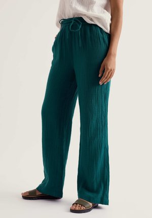 Femeie purtând pantaloni largi, texturizați, de culoare teal, cu talie elastică și cordon, bluză albă lejeră și sandale olive tip slide.