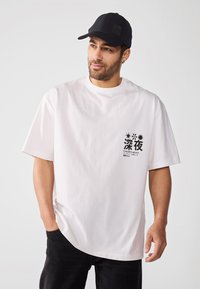 Camiseta de algodón blanca de manga corta, con un gráfico impreso en negro en el área del pecho izquierdo y un corte relajado. Combinada con una gorra negra y jeans oscuros.