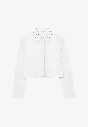 Maglietta bianca corta con maniche lunghe e colletto a punta, caratterizzata da borchie metalliche sul colletto e chiusura frontale con bottoni.