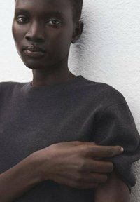 Ciemnoszara bluza z okrągłym dekoltem, z krótkimi rękawami i gładką teksturą, noszona przez osobę na tle fakturowanego białego tła.