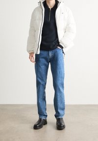 Vit vadderad jacka, svart dragkedja-up tröja, blå raka jeans och svarta läderstövlar. Enkel design med synliga sömmar och dragkedjor.